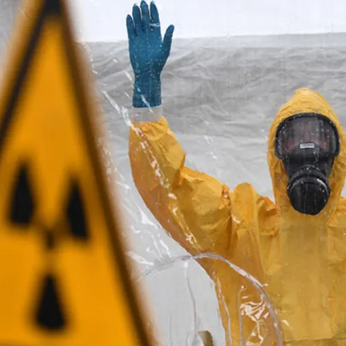 Responder in a yellow protective suit and respirator raising a hand beside a radiation hazard sign during an emergency drill